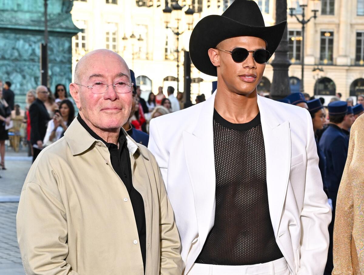 David Geffen, in tan shirt, and Donovan Michaels, in white suit and cowboy hat, stand together on a city street.