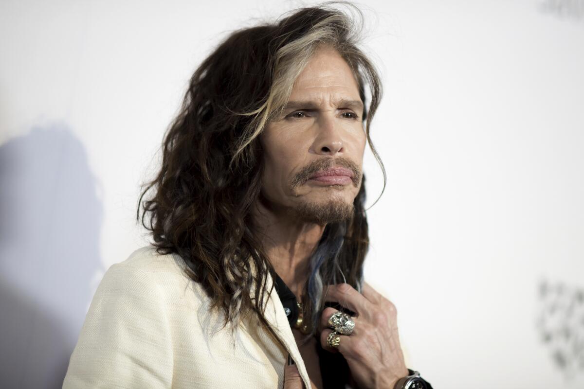 Steven Tyler squints while touching his long hair with one hand and wearing a cream suit jacke