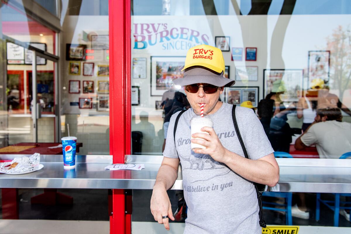 A portrait of a man sipping a milkshake at the outdoor standing counter. He wears a yellow Irv's cap over his other hat.