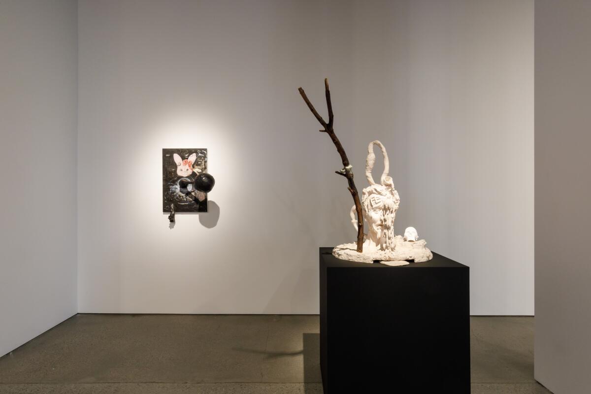 A sculptural artwork on a pedestal and an assemblage piece hung on a blank white wall are spotlighted in a gallery.