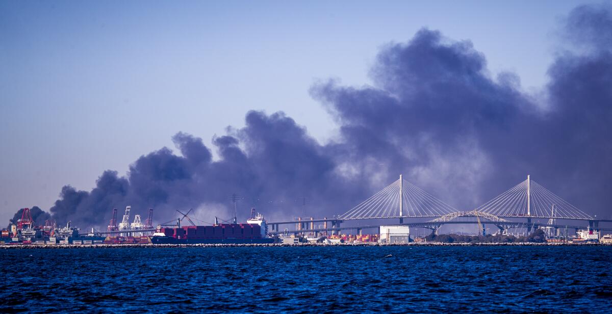 A large, drifting plume of black smoke rises behind a waterfront area