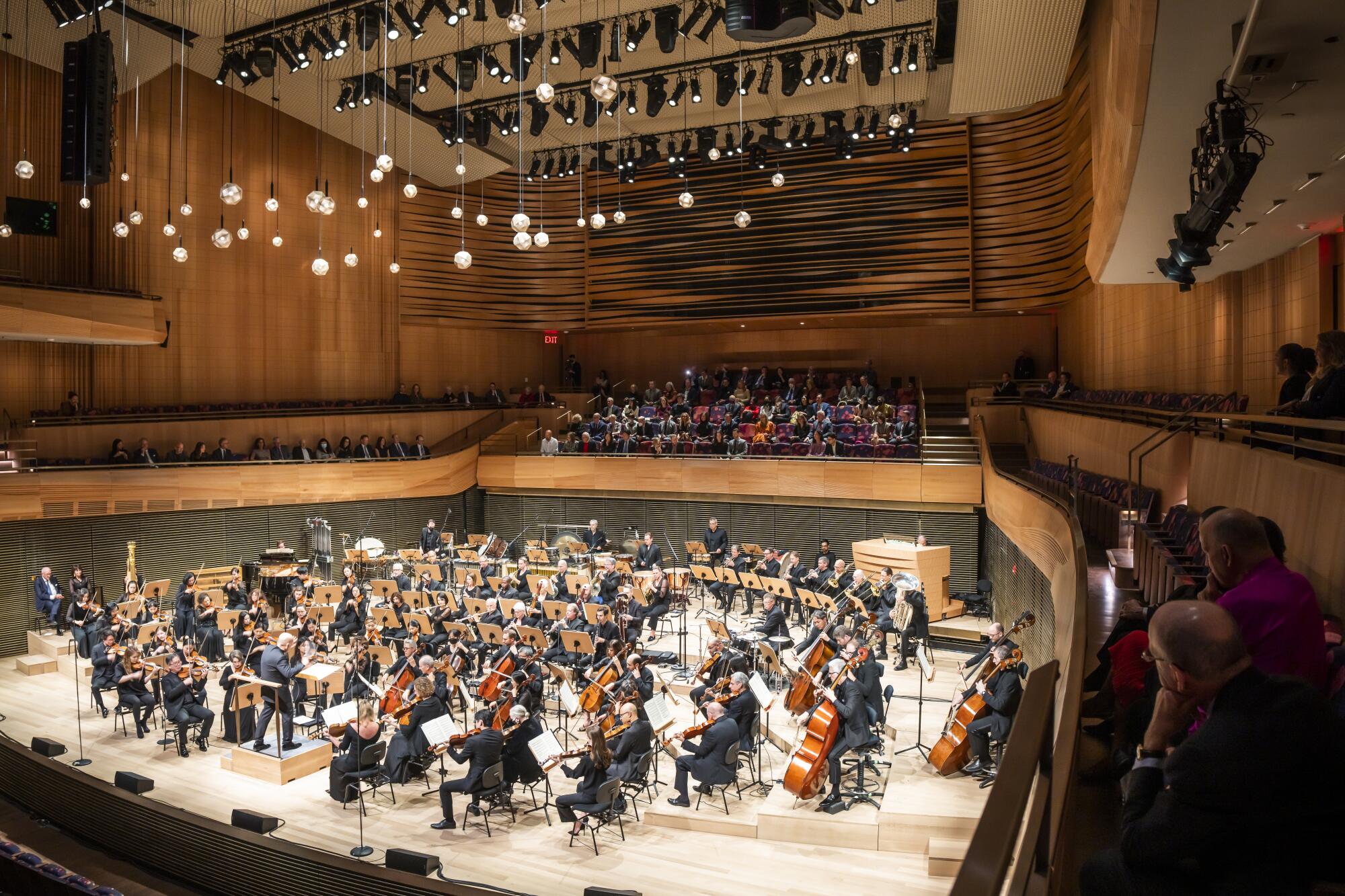An audience is seen surrounding the wooden stage where the New York Philharmonic performs in a wooden concert hall.