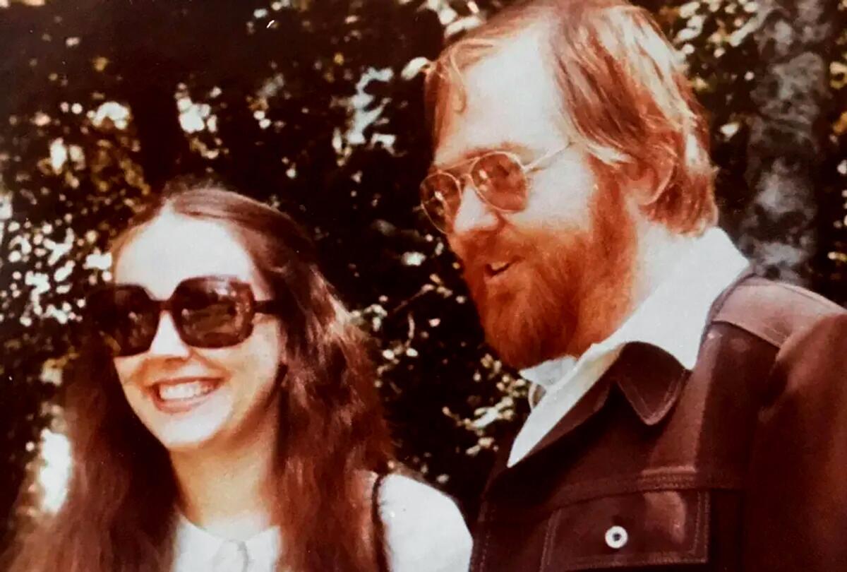 A smiling woman in sunglasses and long hair standing with a bearded, smiling man in sunglasses.