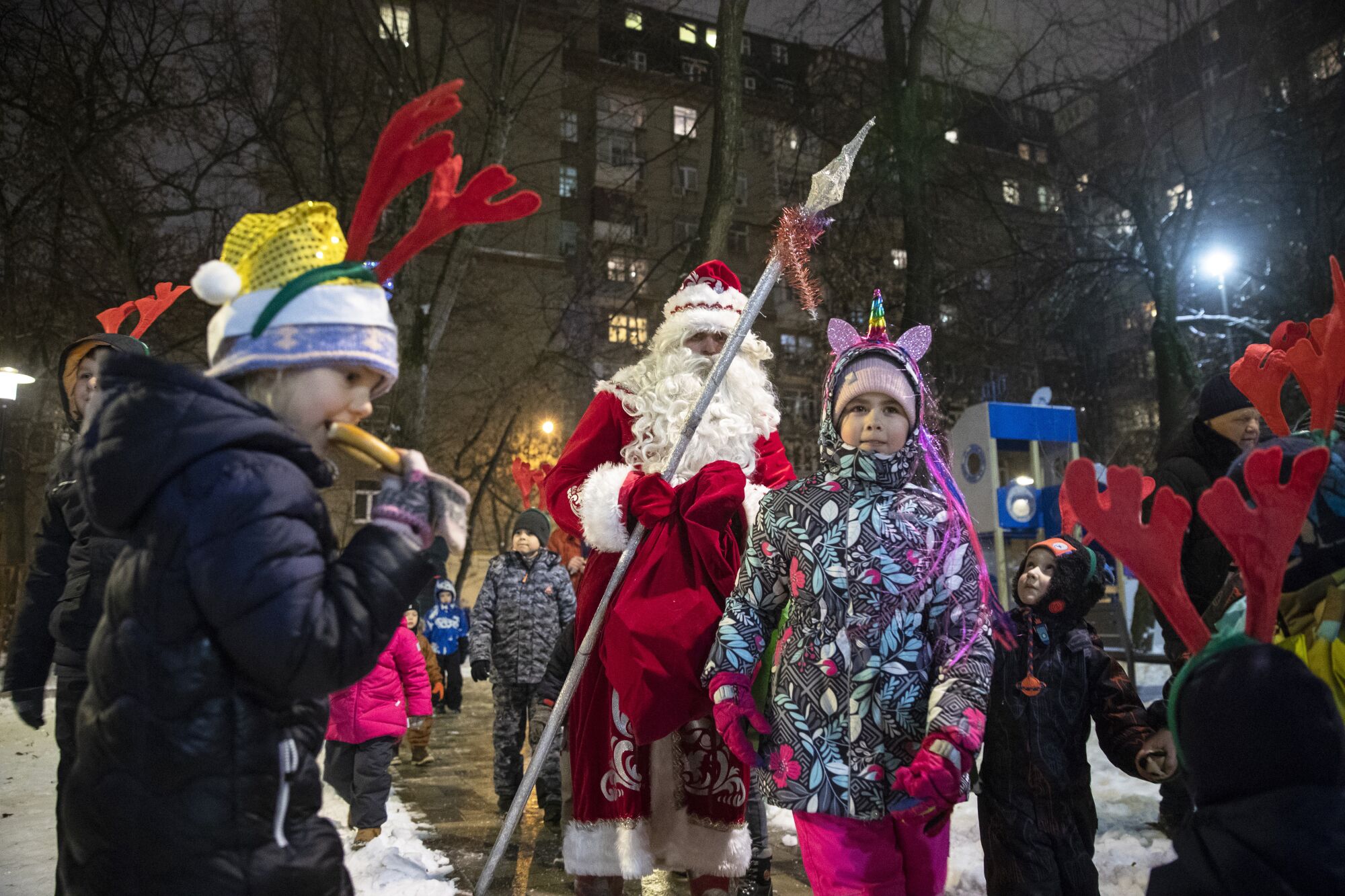 A boy eats a soft pretzel and a girl wears a unicorn headband near a man dressed as Santa Claus.
