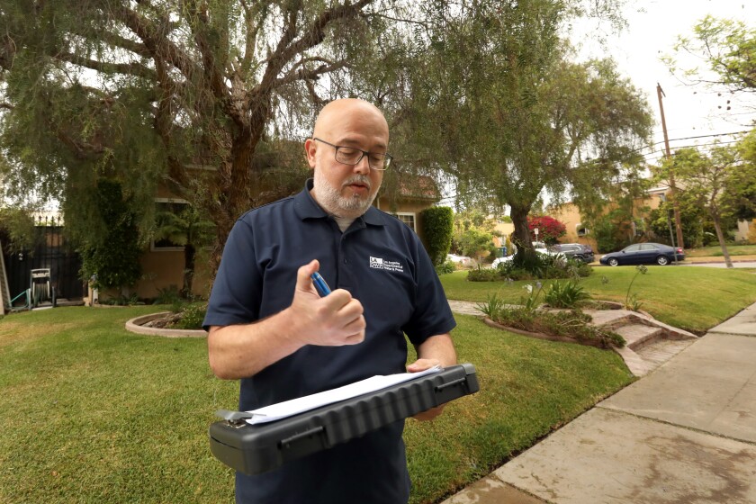 A man with a gray beard and glasses holds a pen in one hand and a clipboard in the other near a front lawn 