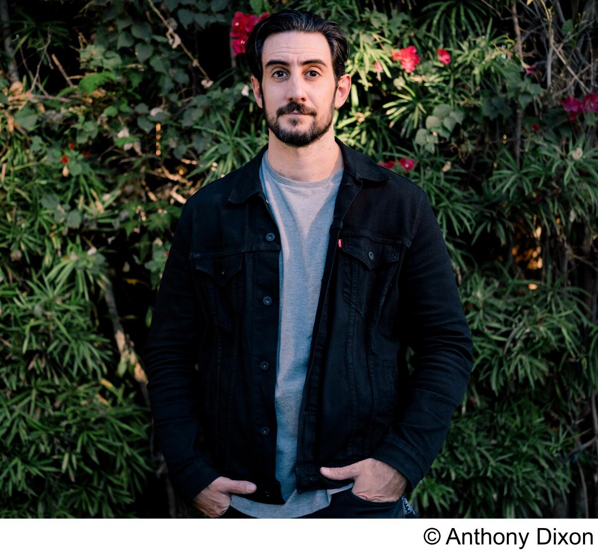 A bearded man in a T-shirt and jacket stands in front of a wall of hedges.