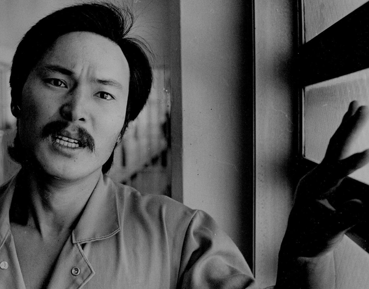 A black-and-white photo of a mustached man who looks angry, standing next to a window.