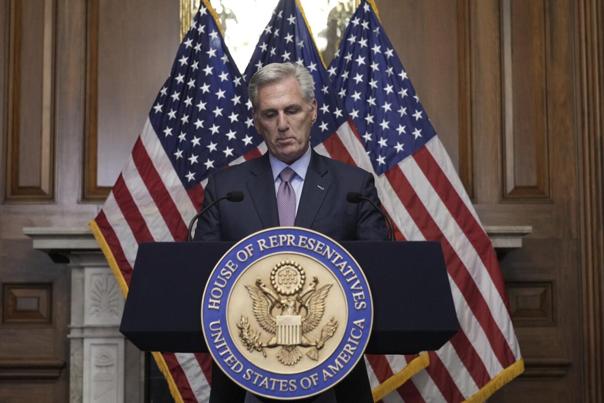 A man with gray hair, in a dark suit, stands in front of U.S. flags at a lectern with a seal of the House of Representatives 