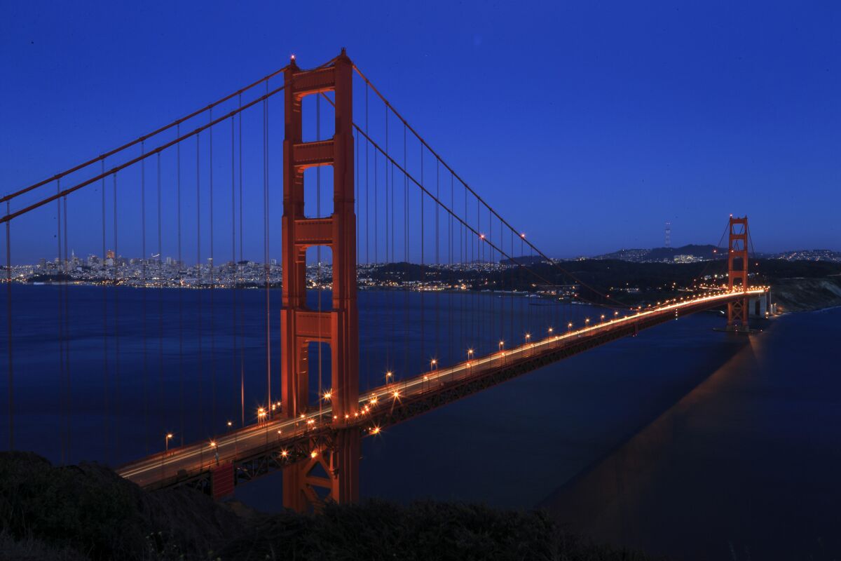 The roadway is lighted on the Golden Gate Bridge at dusk with the shoreline and city in the background.