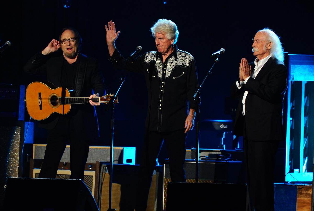 Stephen Stills holding a guitar, left, Graham Nash waving and David Crosby touching his hands together.