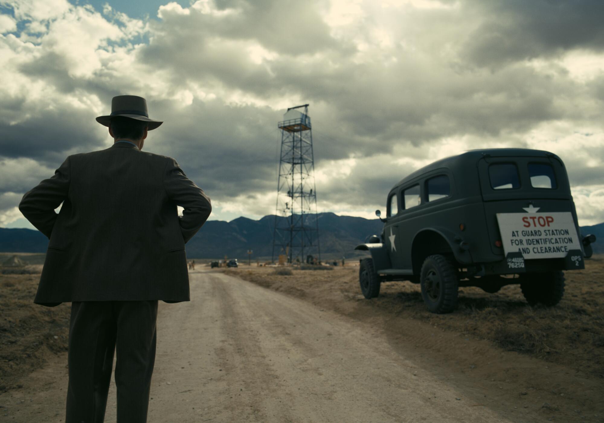 A man in a hat, seen from behind, watches a distant tower from a road, along which is parked a vintage truck.