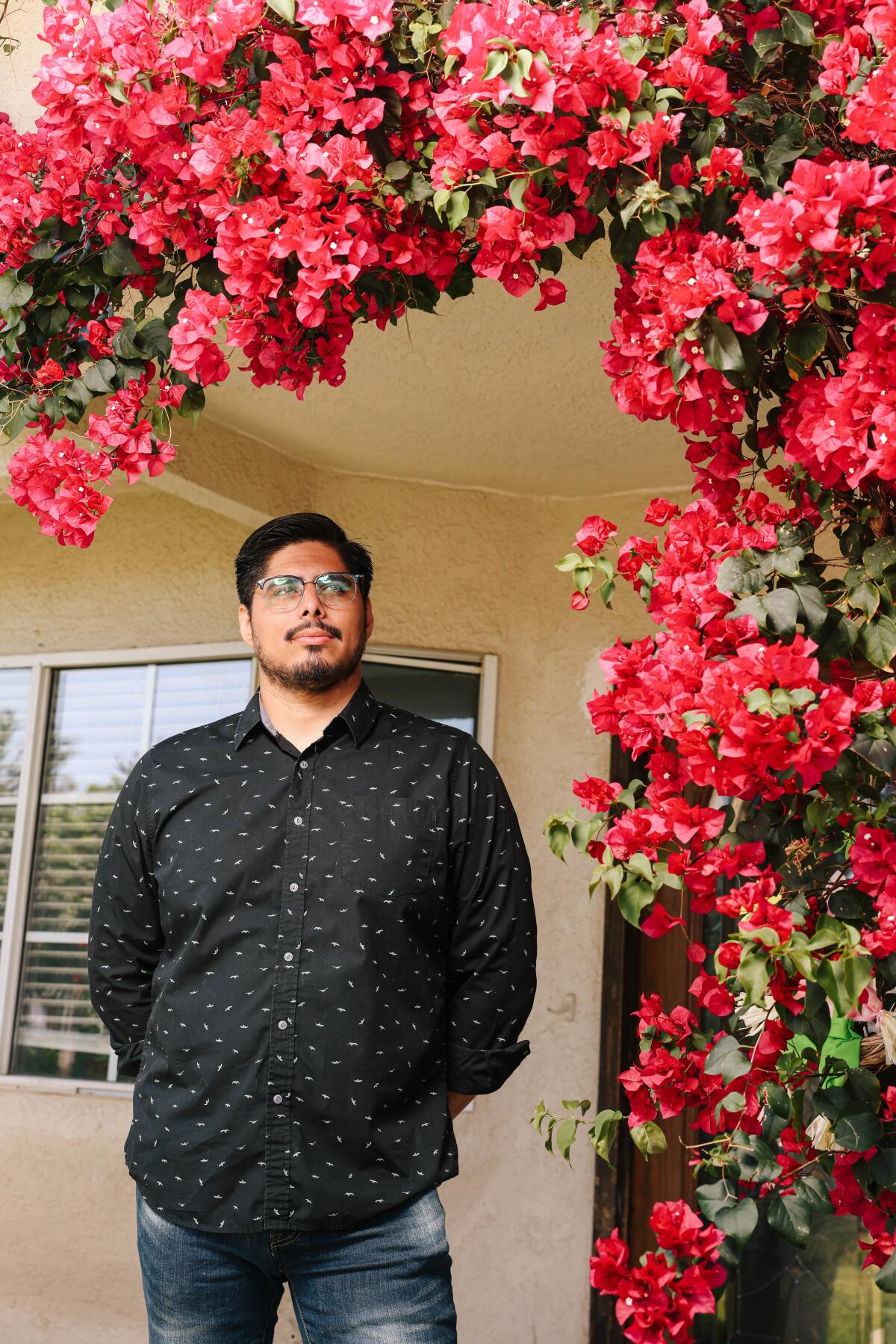 A bearded man in glasses and a black shirt poses amid red blossoms. 