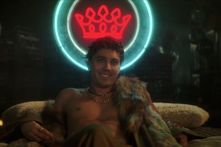 a shirtless Lukas Gage lounging on a bed lit by a sign shaped like a crown
