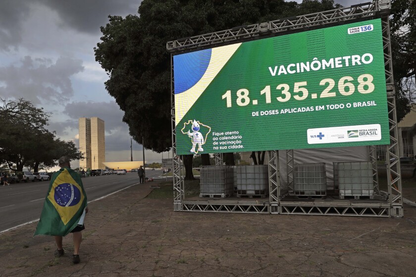 A demonstrator wearing a Brazilian national flag as a cape for a rally in support of Brazil's President Jair Bolsonaro, walks past a billboard presenting the number of COVID-19 vaccines administered to encourage others to get the vaccine, outside the Health Ministry in Brasilia, Brazil, Tuesday, March 30, 2021. (AP Photo/Eraldo Peres)