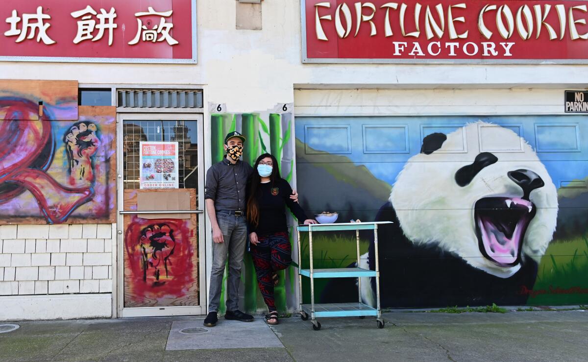 A couple stand in front of their storefront with paintings of raised fists and a panda under a "Fortune Cookie Factory" sign
