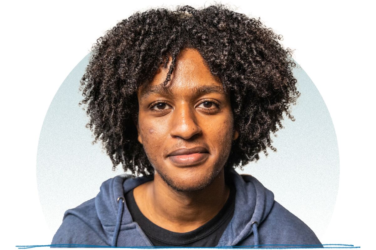 A young man with long black hair in twists wearing a blue hoodie smiling for a portrait.