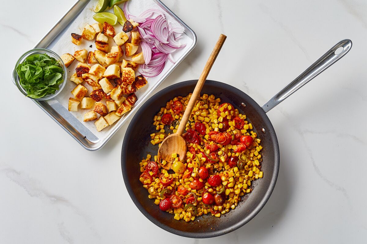 Sauteed corn and cherry tomatoes in a pan, with a tray of halloumi, basil, red onion and lime to be added.
