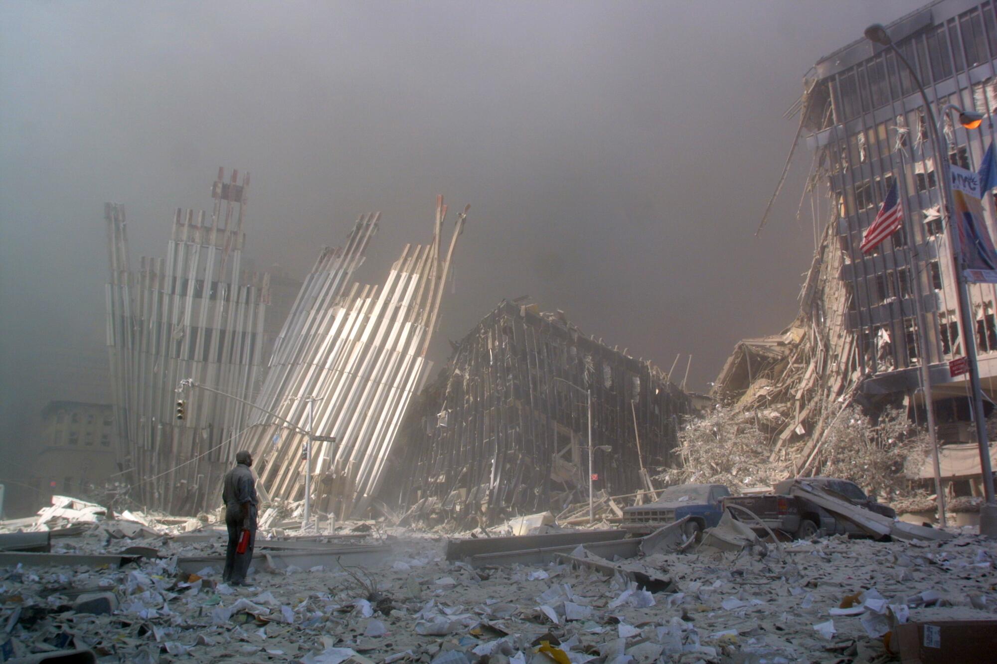 A man stands surrounded by debris, with an American flag on a pole nearby