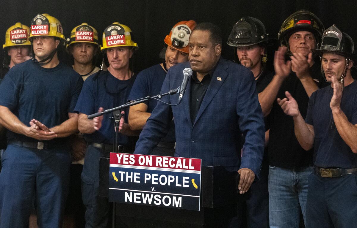 Larry Elder stands before a microphone with firefighters behind him 