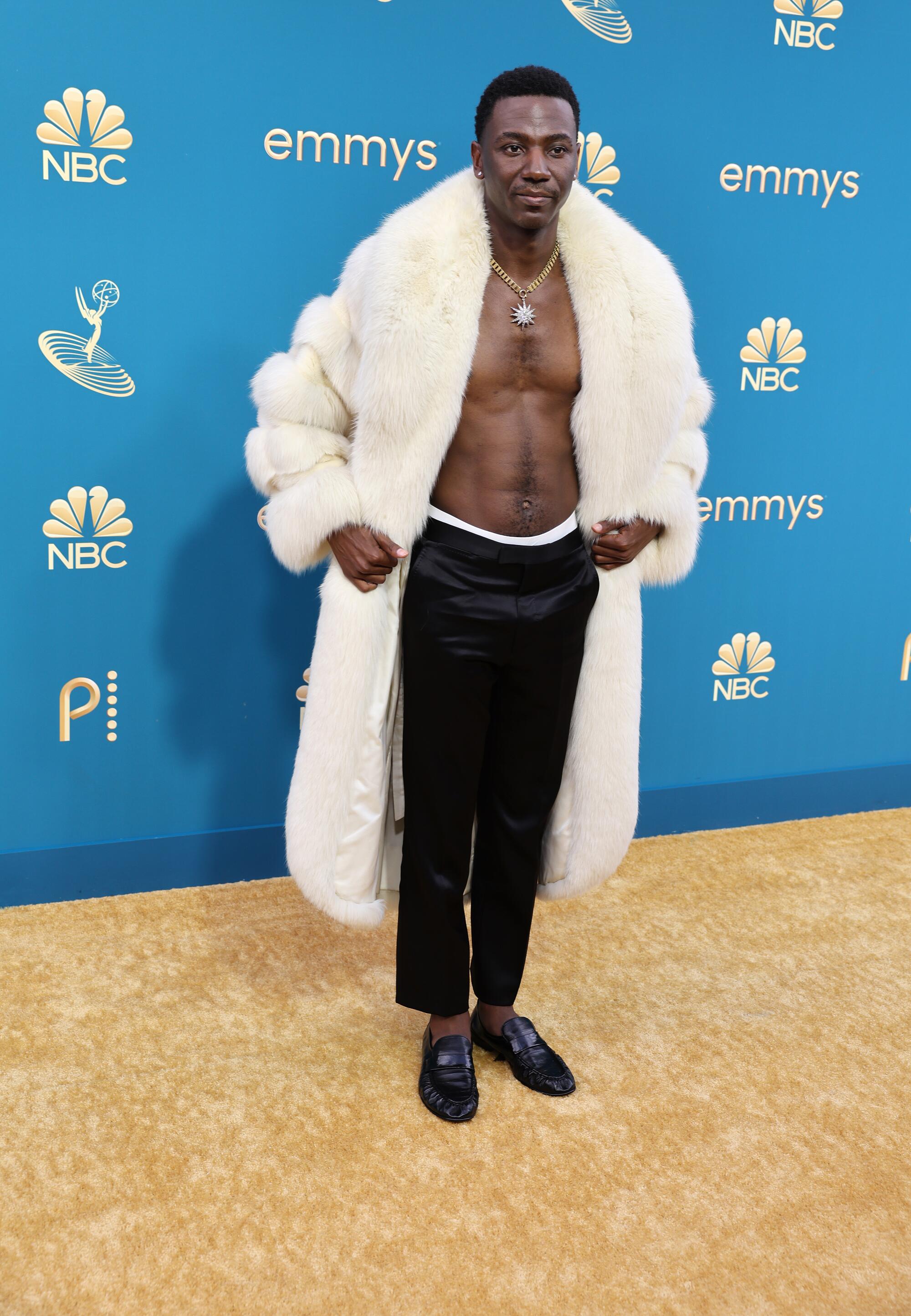 A man in a white fur coat and no shirt walks the red carpet at an awards show