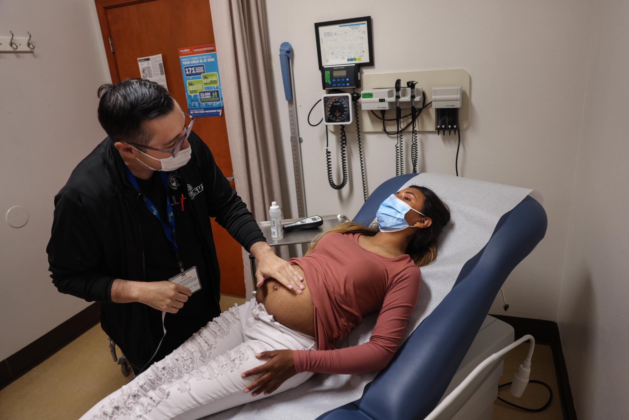 A dark-haired man with glasses touches the exposed belly of a pregnant woman lying in an examination chair 