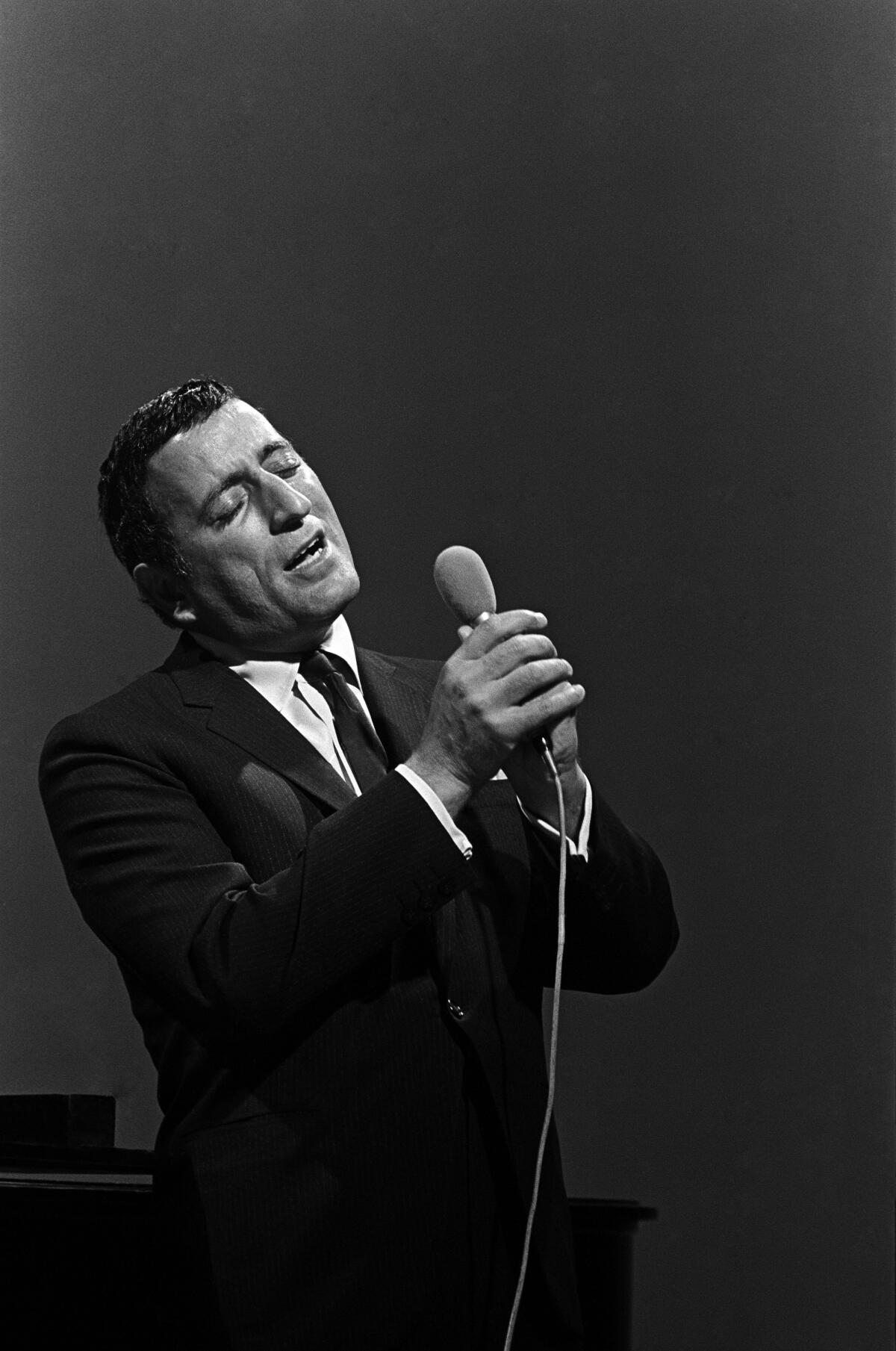 A vertical, black-and-white frame of Tony Bennett performing as he sings into a handheld microphone with his eyes closed. 