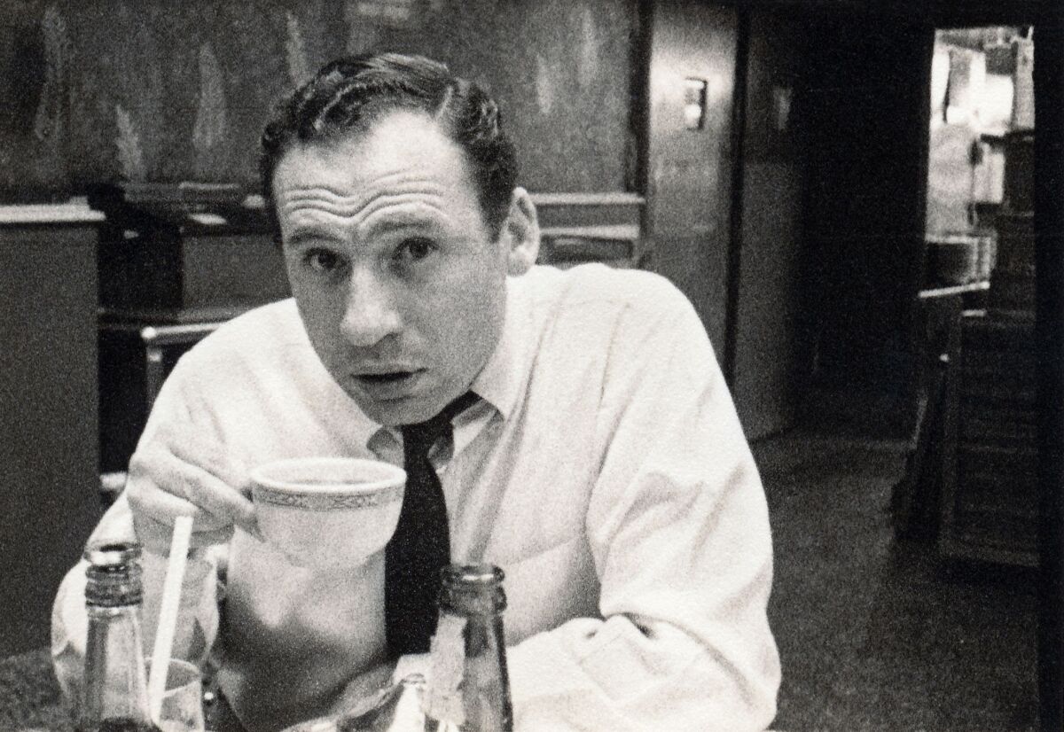 A young Mel Brooks in a white shirt and narrow tie, sitting at a cafe table drinking coffee.