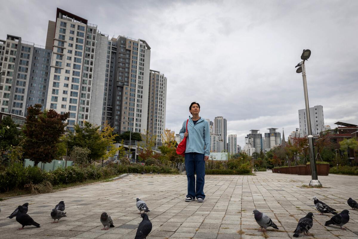 A man in a light blue jacket and dark pants stands holding a red bag near some pigeons, with high-rise buildings behind him