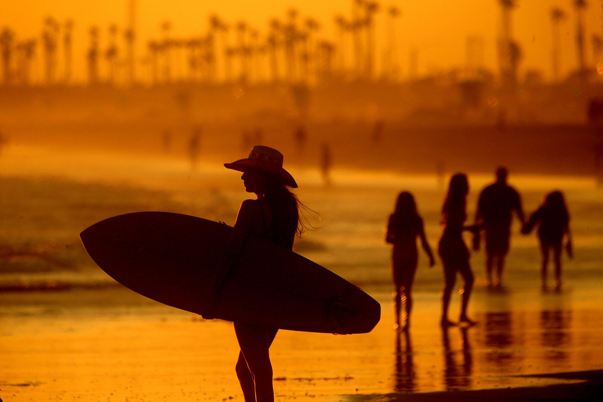 A woman holds a surfboard at a beach that glows orange in the sunset.