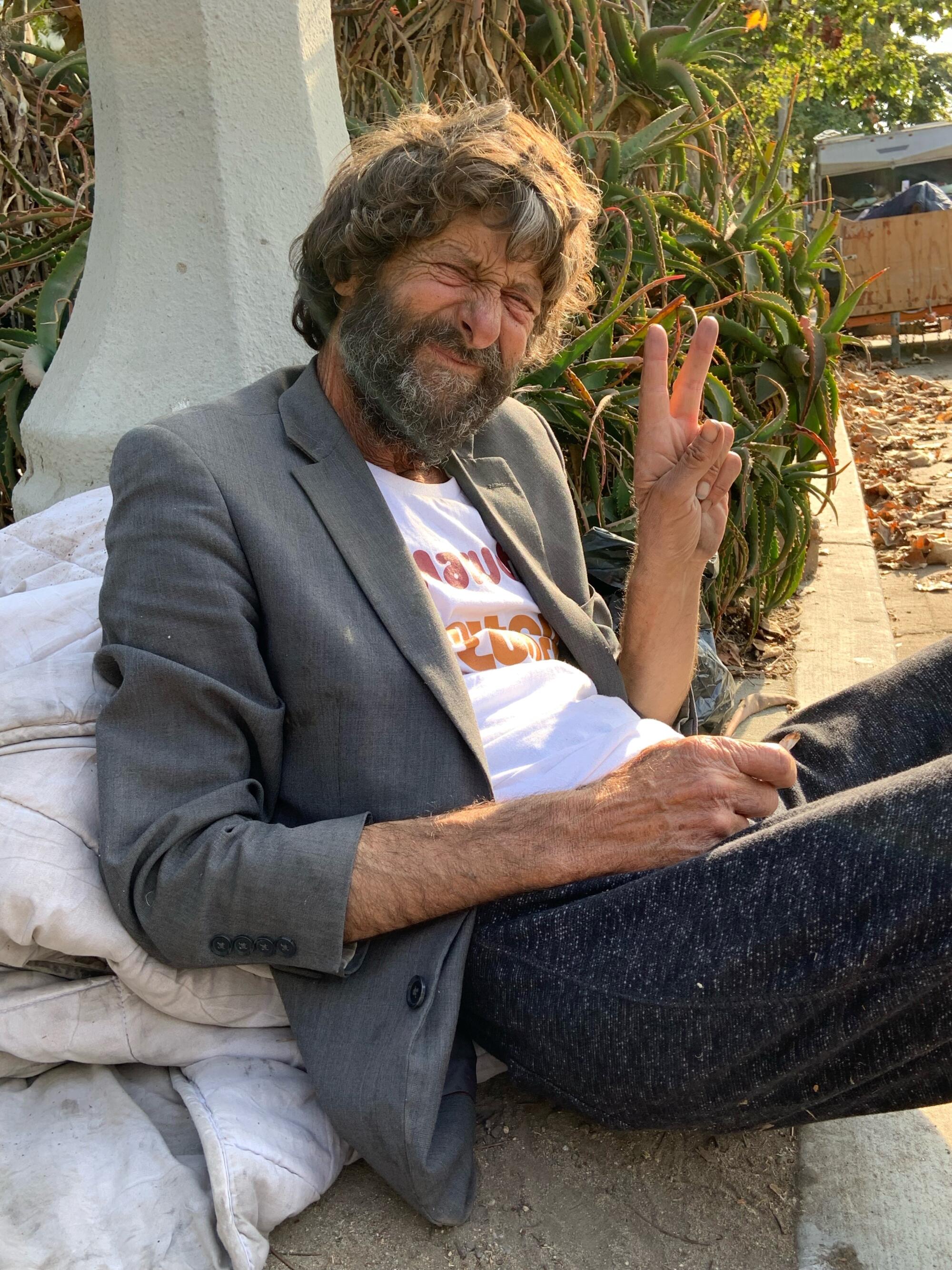 A man in a jacket and T-shirt leans against a comforter while holding up a peace sign