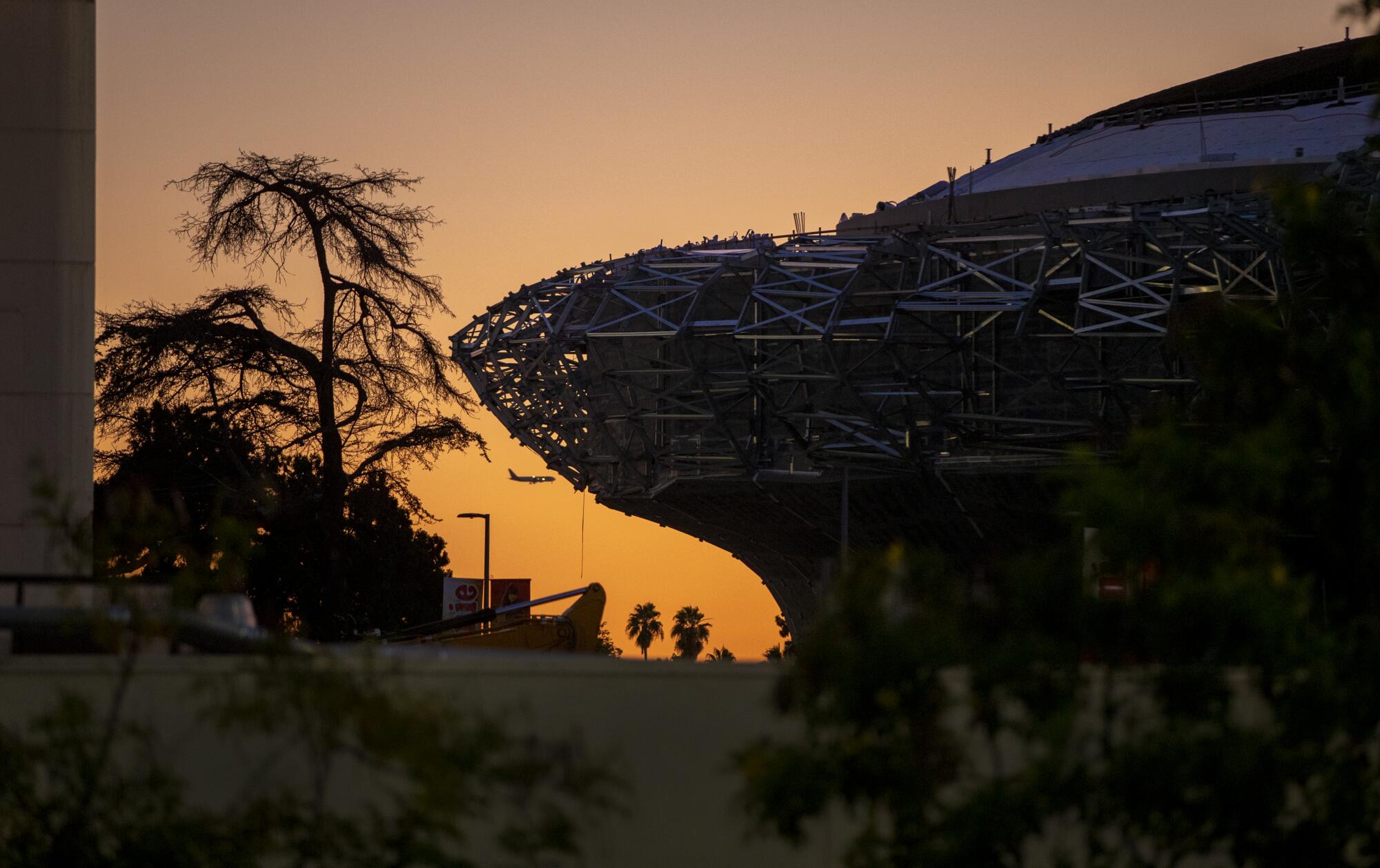 A glowing sky at sunset silhouettes an airplane and the Lucas Museum of Narrative Art under construction in Exposition Park.