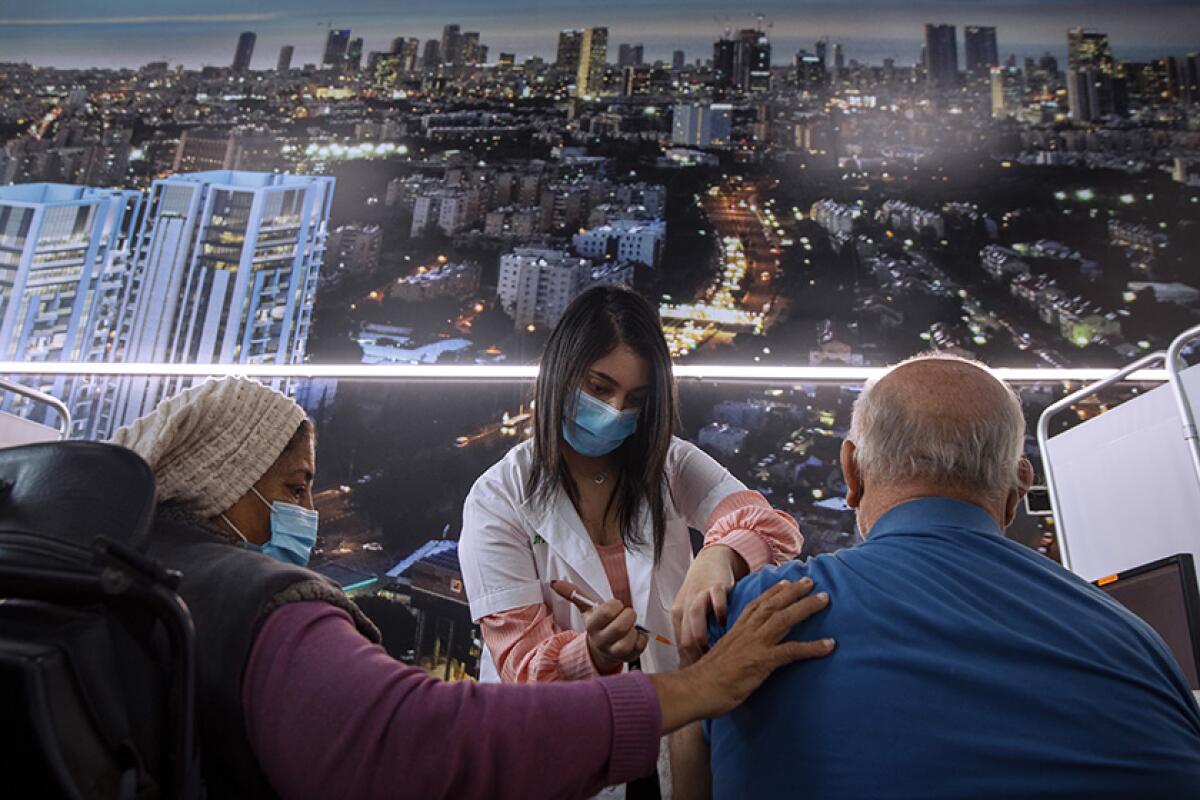 An Israeli man receives a COVID-19 vaccine from a healthcare worker while another woman rests her hand on his shoulder.