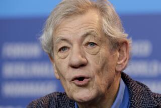 Ian McKellen looking off with a parted mouth wearing a dark coat in front of a blue backdrop