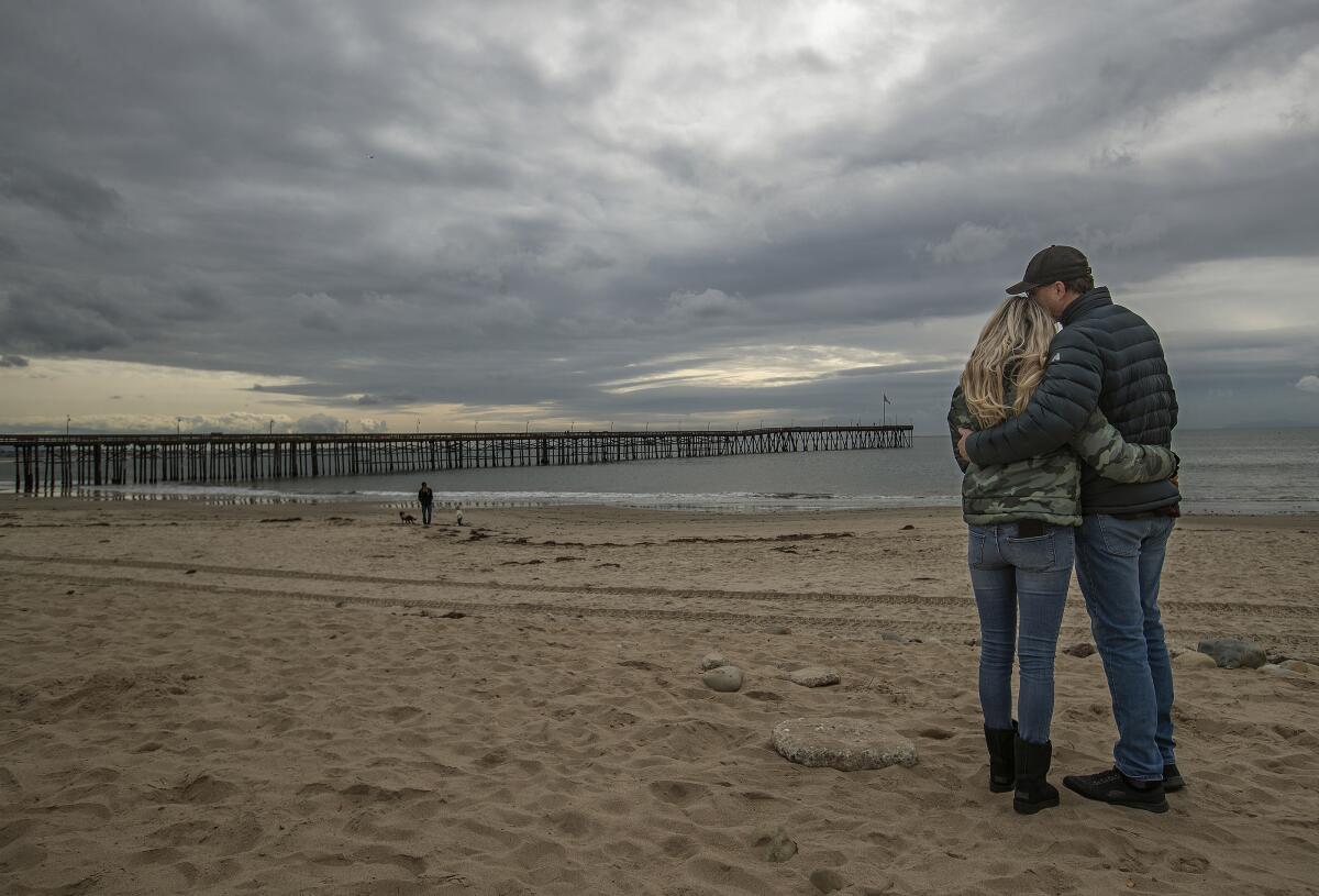 A couple embrace on a beach with a pier under dark, cloudy skies
