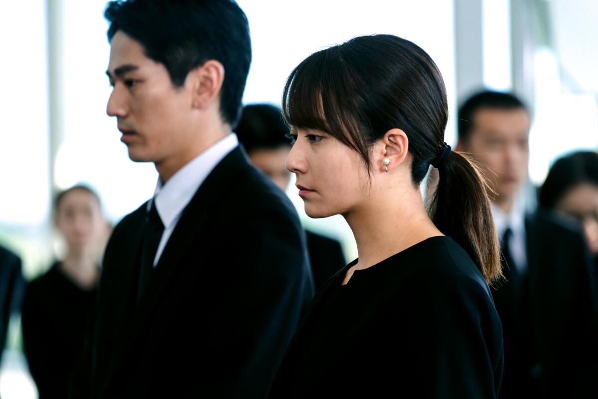 A somber man and woman, both dressed in black, stand together.