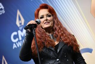 An angled photo of a woman with long, wavy red hair in a black leather jacket standing on a stage holding a microphone