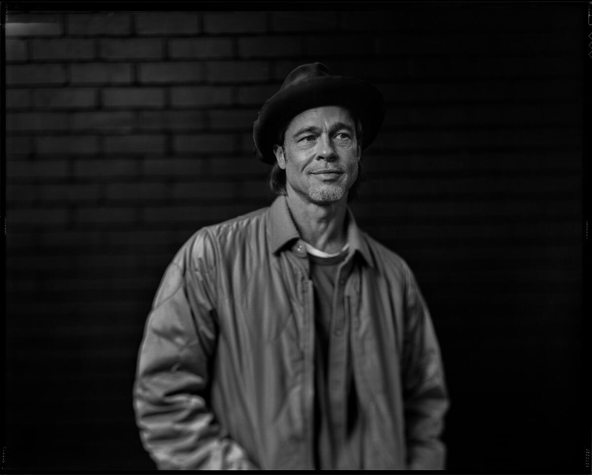 A man in a brown leather jacket and fedora standing in front of brick wall