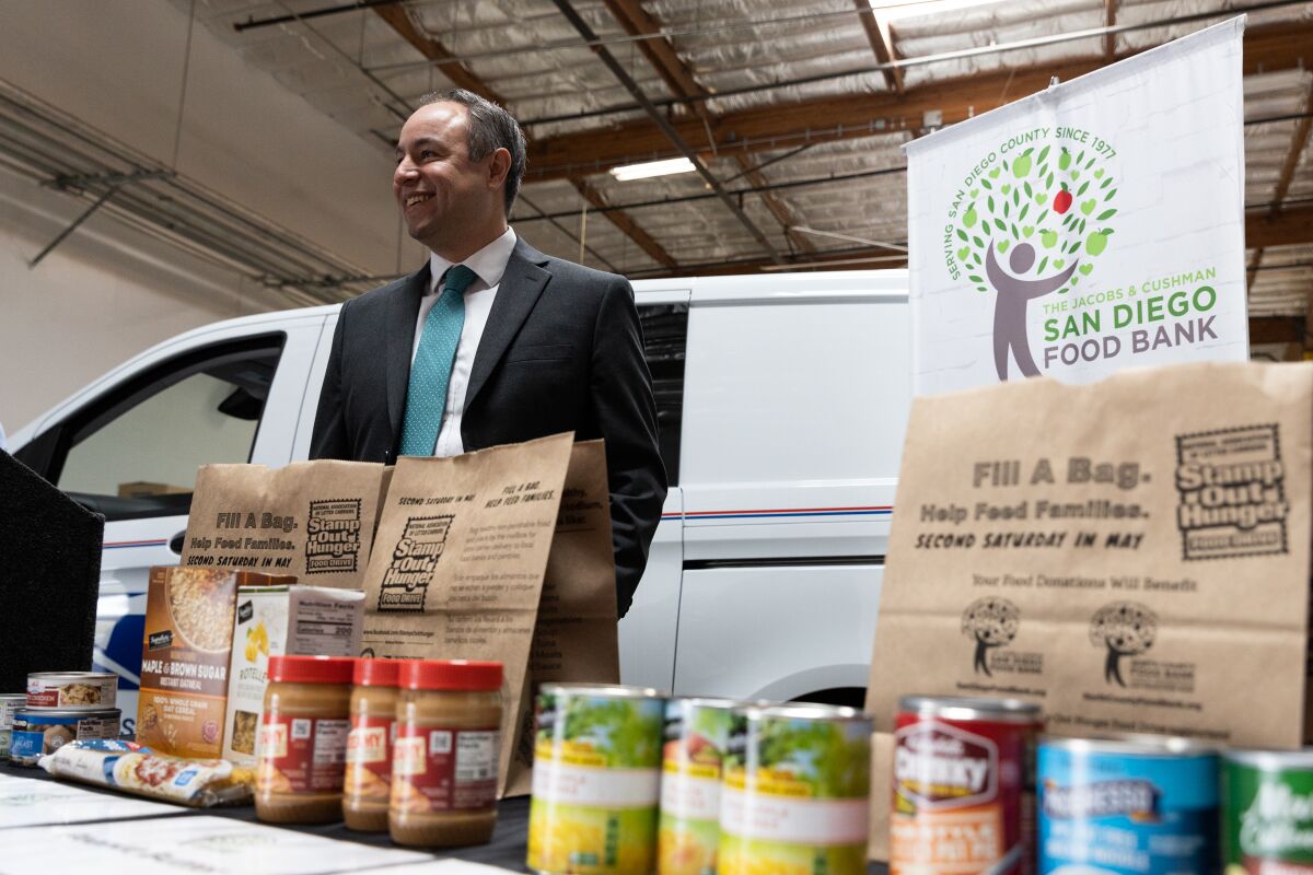 A man in a suit smiles as he stands in a food bank warehouse behind a display of canned food on a table.