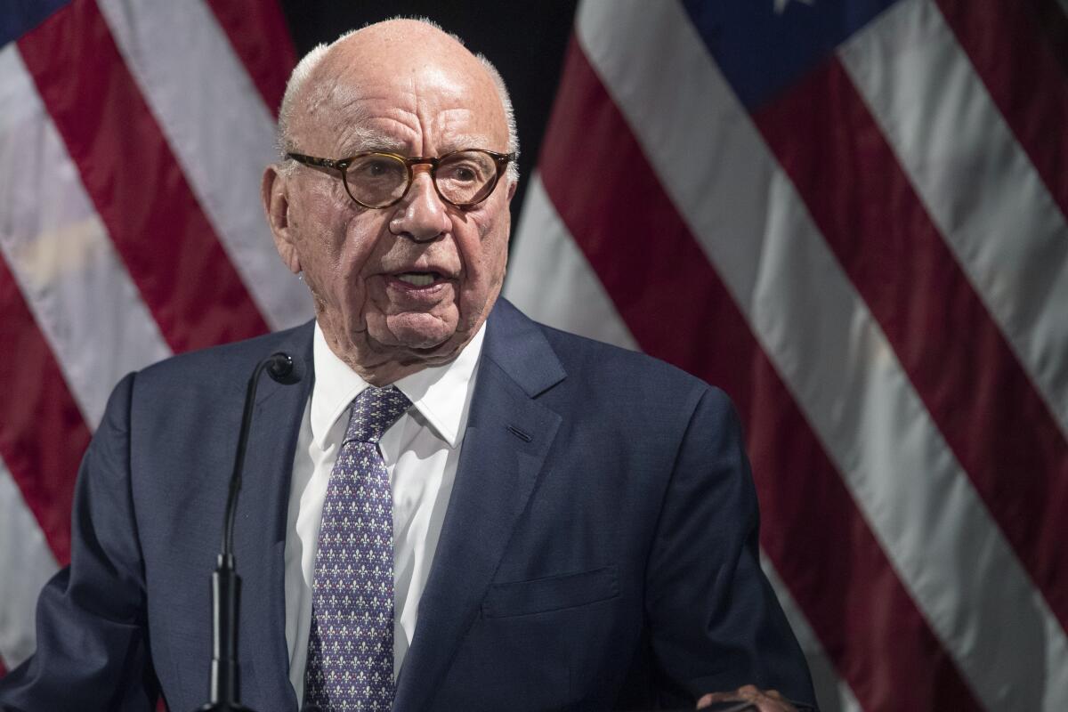 Wearing a suit, Rupert Murdoch stands in front of American flags