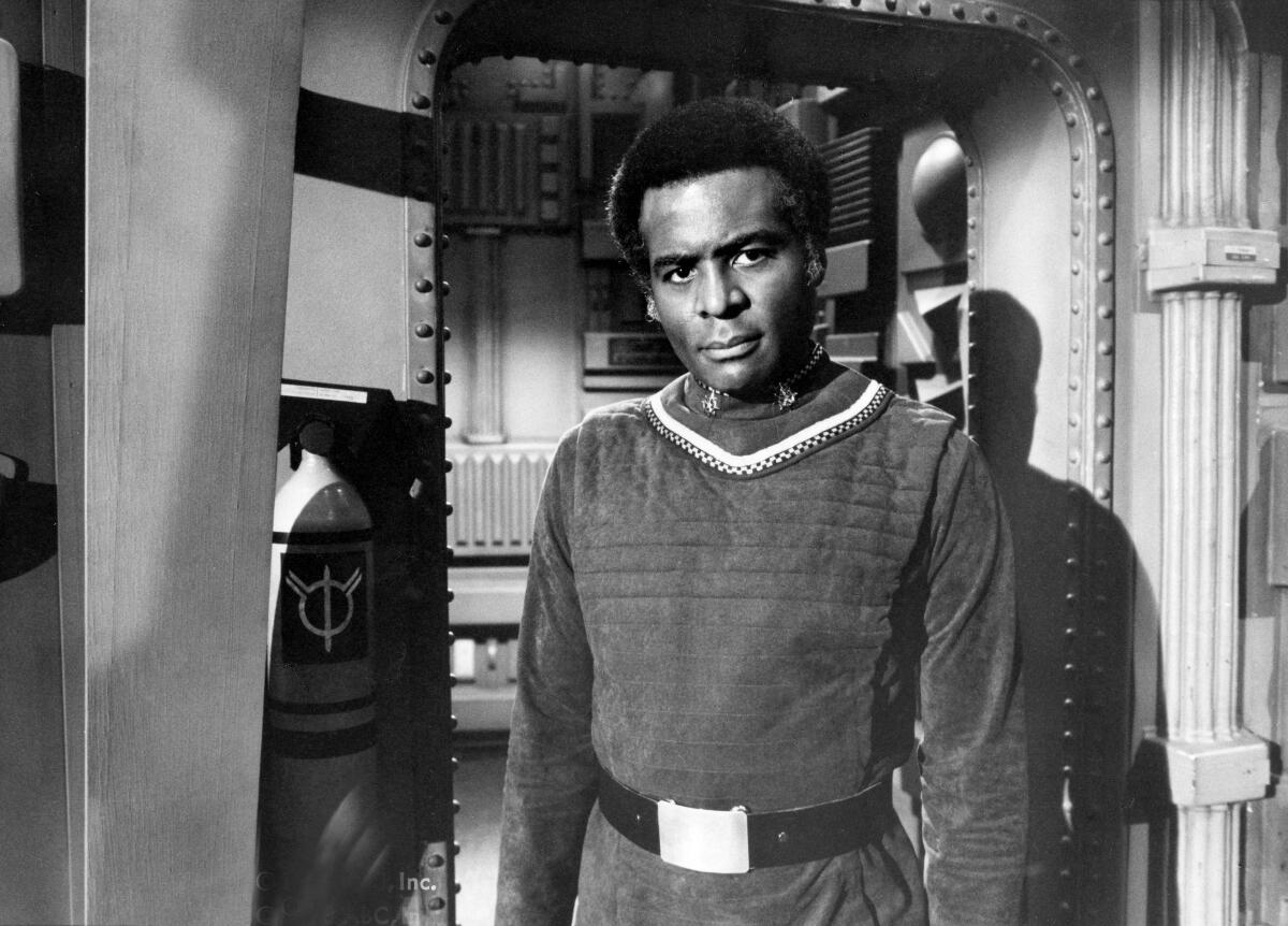 A black-and-white photo of a Black man in a tunic with a wide belt standing and looking sternly ahead of him