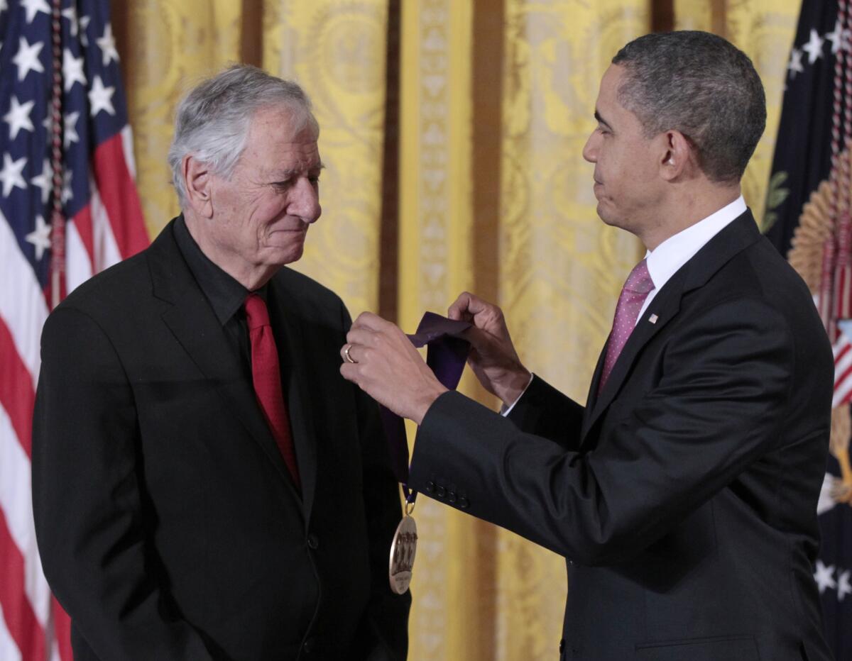 Robert Brustein, an older white man, stands before a U.S. flag as President Obama prepares to  present him with a medal