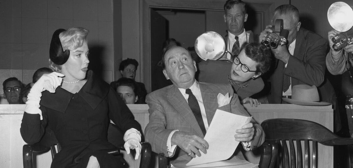 Marilyn Monroe sits alongside her attorney in court alongside photographers holding flash cameras. 