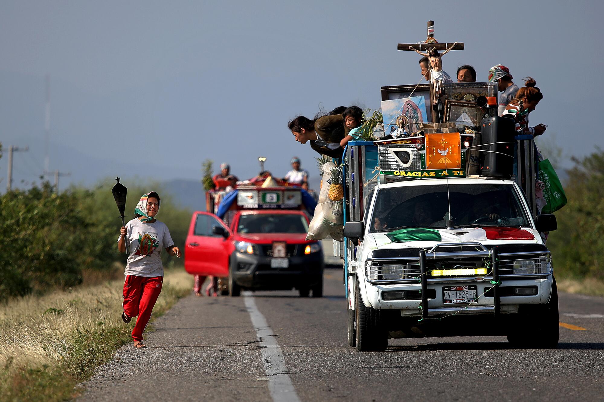 A woman runs alongside a procession of vehicles with a crucifix.