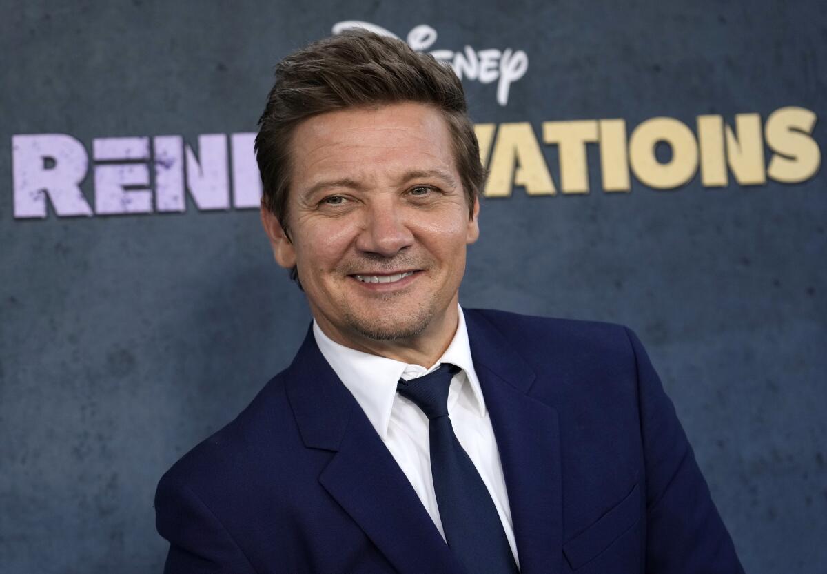 Jeremy Renner smiles and looks to the side in a blue suit and tie