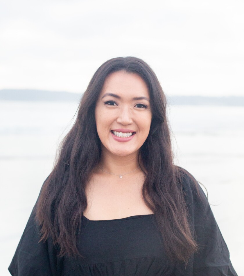 photo of a woman with long, black, wavy hair wearing a black, long-sleeved blouse and standing in front of the ocean