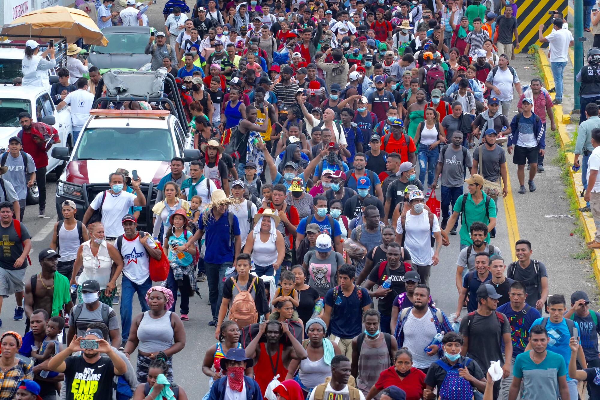 Crowds of people dressed in short sleeves or tank tops, many wearing backpacks, walk on a road alongside vehicles. 