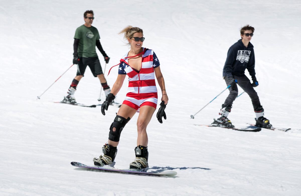A woman at the center in summer outfit snowboards down a shallow hill. Two skiers without coats are riding behind.