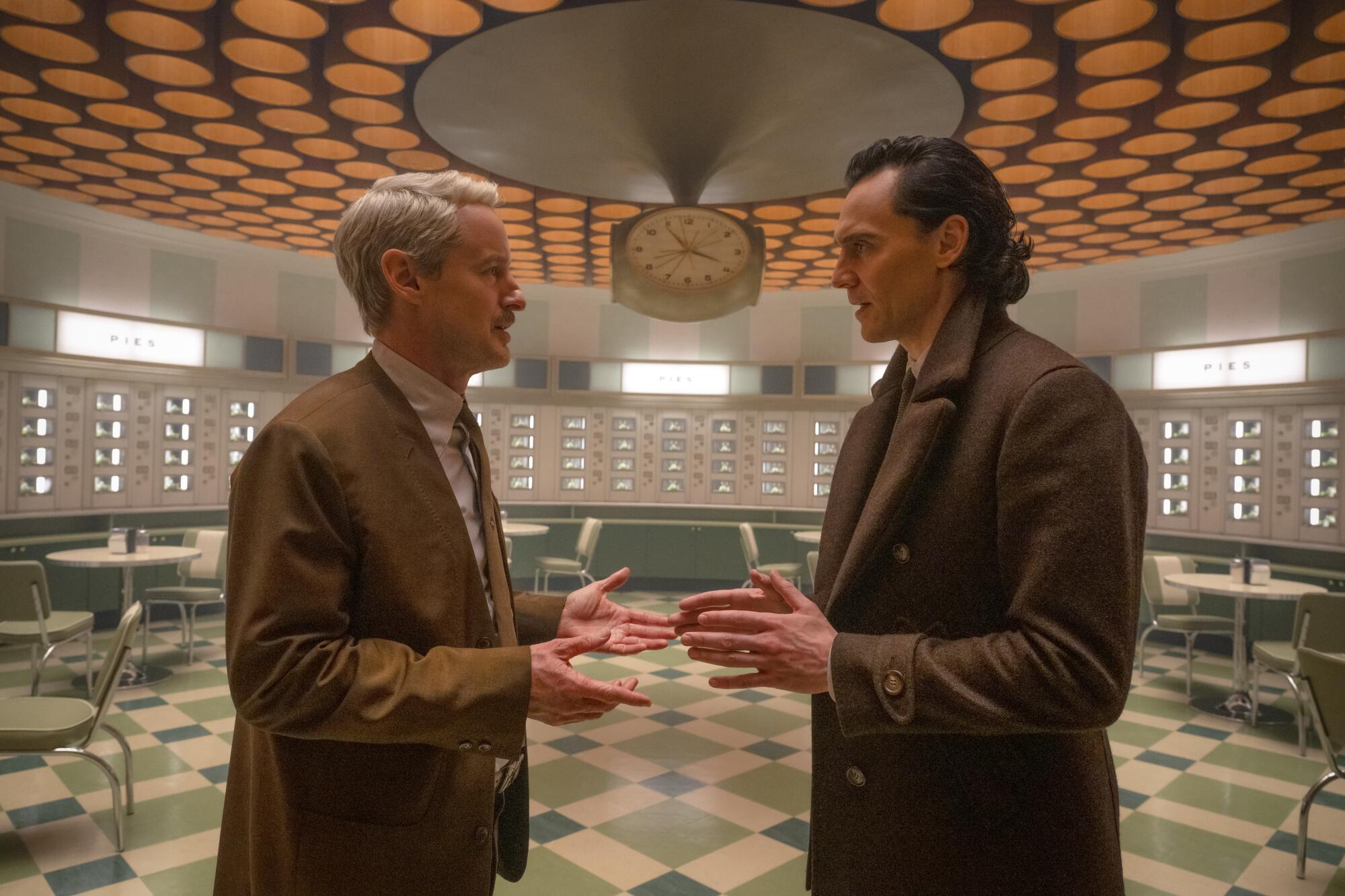 A gray-haired man in a brown suit speaks to Loki in an empty green and white diner.