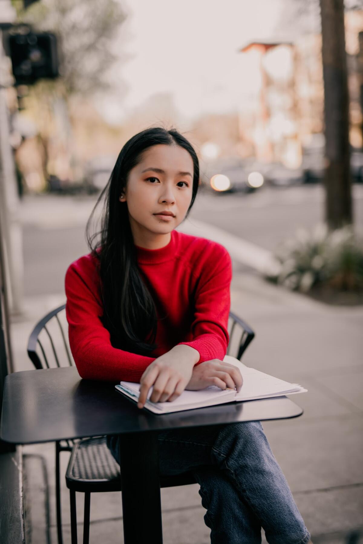 A young woman sits at a cafe table on a sidewalk, hands folded over a notebook.