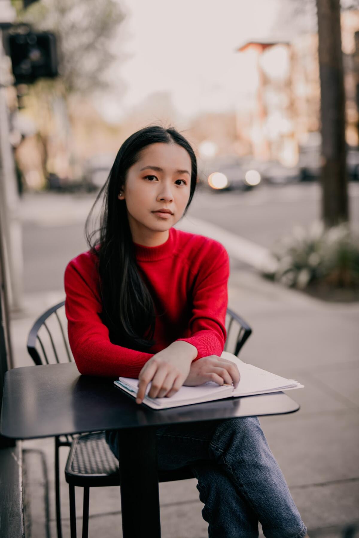 A young woman sits at a cafe table on a sidewalk, hands folded over a notebook.
